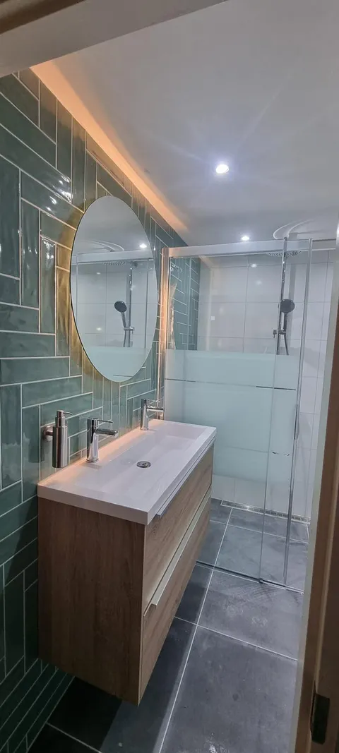 Badkamer met groene tegels in visgraatverband, ronde ledspiegel en houten wastafelmeubel