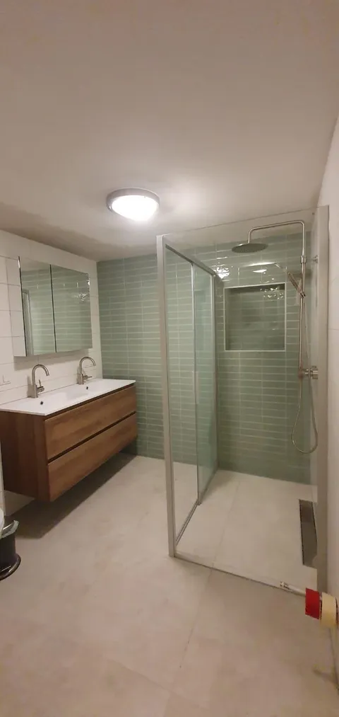Badkamer met groene wandtegels in de douche, glazen douchewand en houten wastafelmeubel