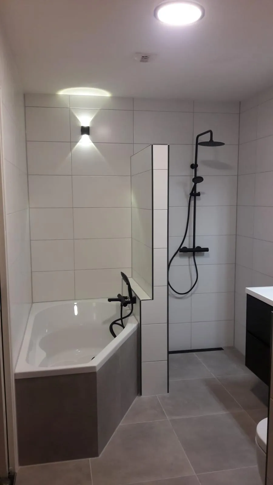 Badkamer met hoekbad, betegelde tussenwand en inloopdouche met zwarte regendouche