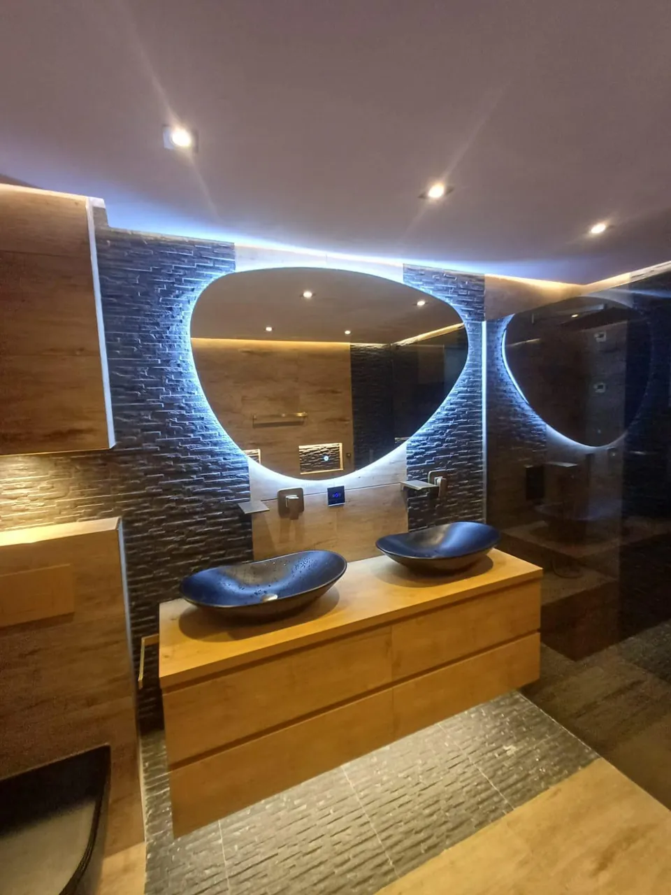 Badkamer met verlichte spiegel, twee zwarte waskommen op een houten meubel en een wand van natuursteenstrips