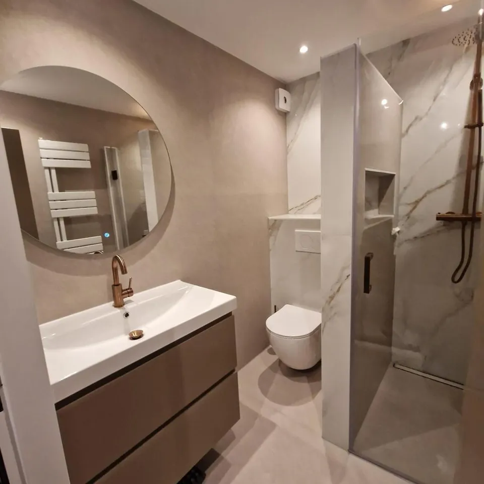 Badkamer in marmerlook met ronde spiegel, wastafelmeubel, hangtoilet en inloopdouche met koperen kraan