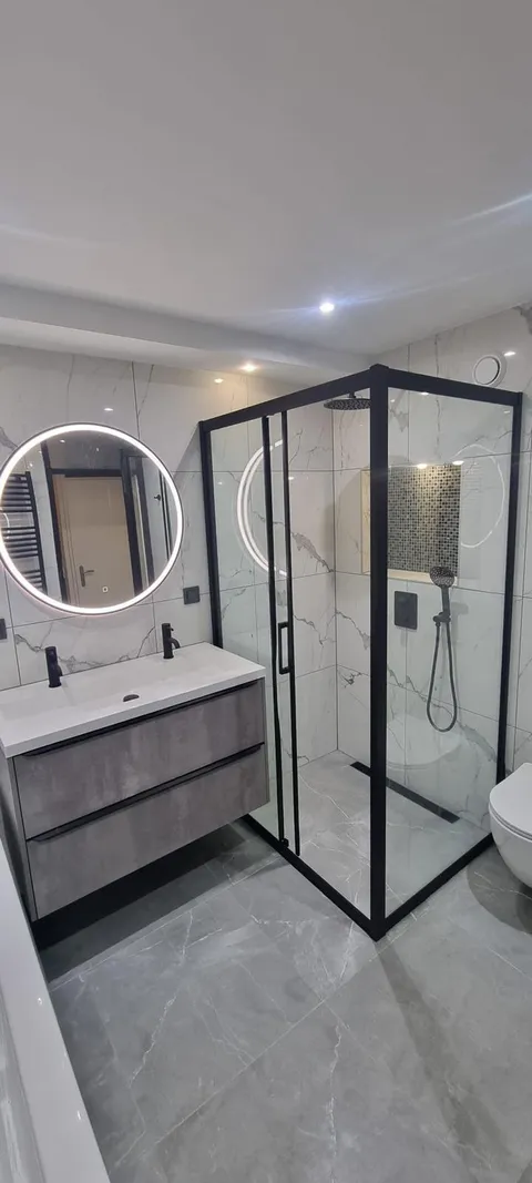 Badkamer met ronde ledspiegel, wastafelmeubel en douchecabine met zwart kader