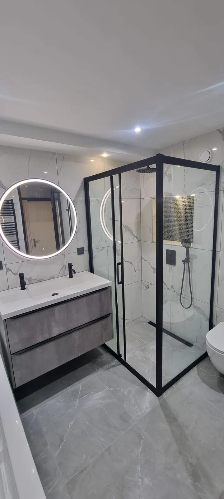 Badkamer met ronde ledspiegel, wastafelmeubel en douchecabine met zwart kader