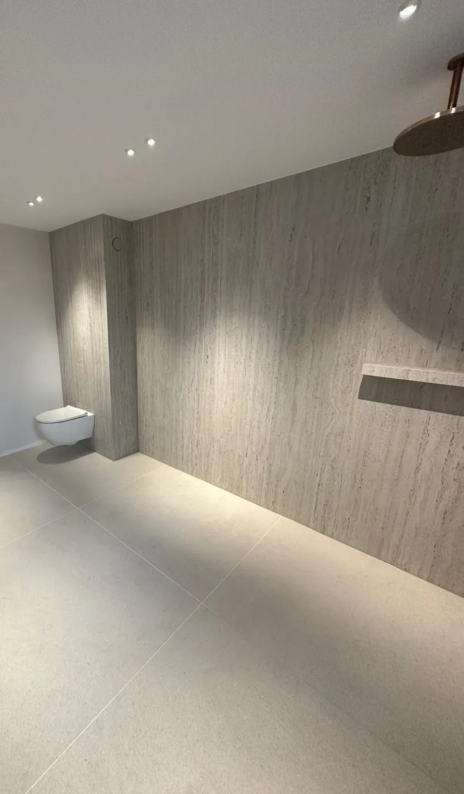 Ruime inloopbadkamer met travertinlook wand, inbouw regendouche en hangend toilet